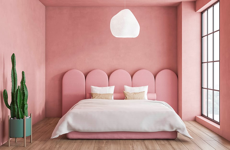 Bedroom in pink, with pink walls, bed with a bed frame in the same colour and white sheets. The headboard of the bed is pink, formed by several aligned arched elements. An irregularly shaped white chandelier descends from the ceiling and a large window illuminates the room from the right. The floor is parquet and there is a large cactus in a vase;