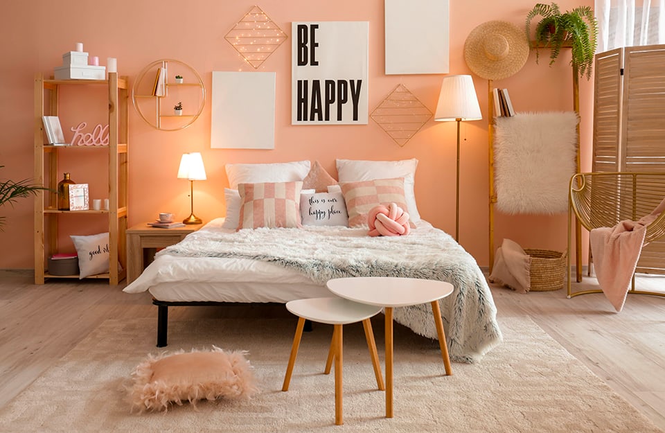 Rustic-romantic style bedroom with pink walls and light parquet flooring. Under the bed is a large cream-coloured carpet. The bed has a simple black metal frame and there are lots of pink cushions with writing on them. At the end of the bed is a pink furry pillow and a pair of irregularly shaped white coffee tables. Behind the bed some prints and a metal grid with small LED lights. On the same wall, to the left of the bed, a simple wooden coffee table with a lamp on it, a circular metal shelf hanging on the wall, and a simple wooden bookcase with various objects on display. On the other side, a floor lamp, a rung ladder holding a furry rug and books. There is a straw hat hanging, and a wooden partition, as well as a bamboo chair