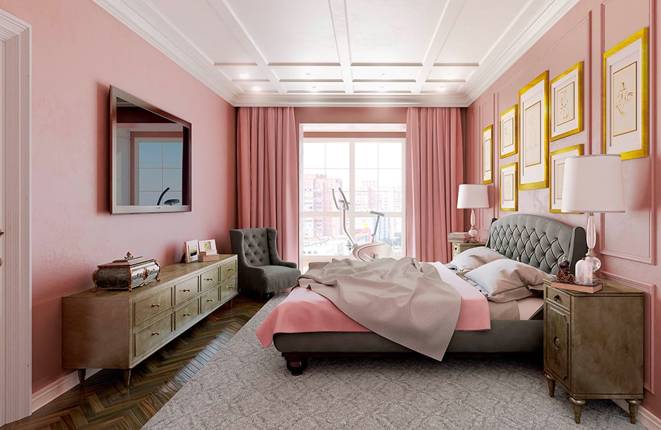 Dormitorio grande con paredes rosas, vista lateral. Las paredes están decoradas con marcos, al igual que el techo, que es blanco. El suelo es de parqué oscuro y, bajo la cama, hay una gran alfombra gris. La cama tiene un marco tapizado en gris y el cabecero también está tapizado en el mismo color. La ropa de cama es gris y rosa. A ambos lados de la cama hay dos mesillas de noche de estilo clásico con lámparas, libros y objetos sobre ellas. En la pared sobre la cama hay algunos cuadros con marcos amarillos. En la pared opuesta hay un gran televisor colgado de la pared y debajo una consola clásica con muchos cajones y cachivaches. Al fondo de la habitación hay una ventana que da a un panorama urbano. Las cortinas son rosas como las paredes y delante de la ventana hay una bicicleta estática;