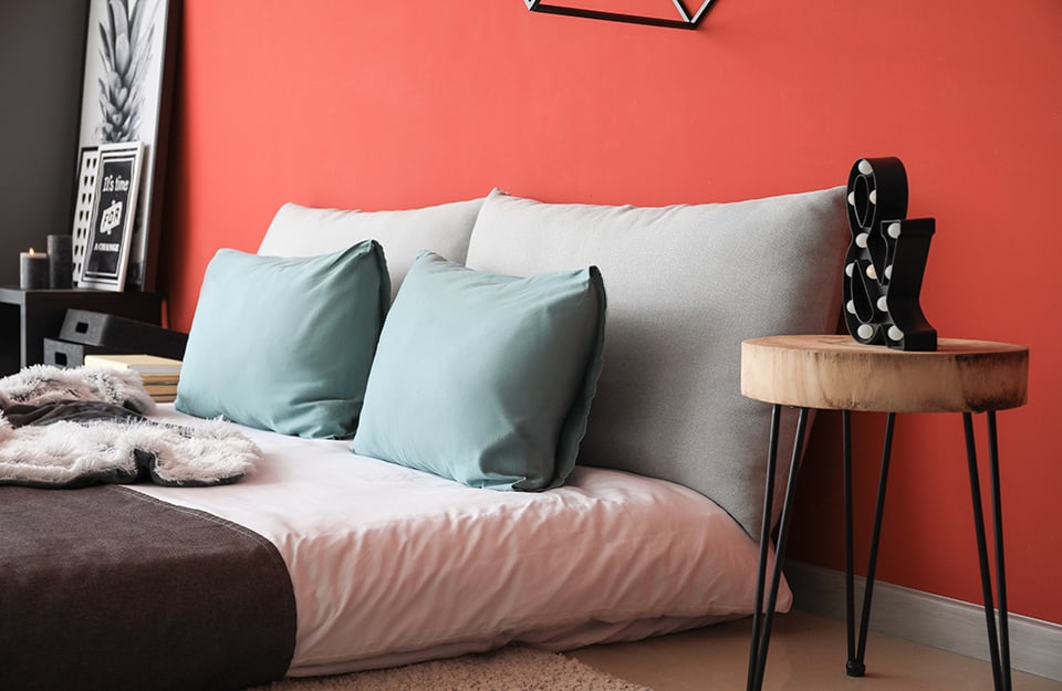 Detalle de una cama con colchón directamente en el suelo, en una habitación con una pared roja. A un lado de la cama hay un taburete con un 