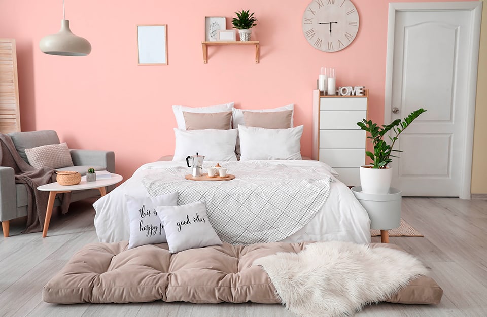 Modern romantic style bedroom with pink walls and light parquet floor. The double bed has white and grey bed linen. At the foot of the bed is a large beige pillow with a blanket and pillows with writing on it. On one side of the bed is an irregularly shaped white table with a wicker basket and a potted plant on top, and a small grey two-seater sofa. On the other, a tall wooden chest of drawers with white drawers and above them candles and letters forming the words 'LOVE'. There is also a small white cylindrical table with a plant in a white pot on top. On the wall is an empty picture frame, a shelf with frames and a plant and a hanging clock. A minimal white chandelier descends from the ceiling. There is a white door to the right