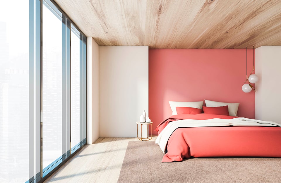 Large bedroom with a fully windowed wall. The décor is minimal, with a wooden ceiling and parquet floor, white walls and a pink recessed wall where the bed is located, with white and pink bed linen. Under the bed is a large beige carpet. To one side of the bed is a circular metal bedside table with white vases on it. Two white spherical lamps descend from the ceiling;