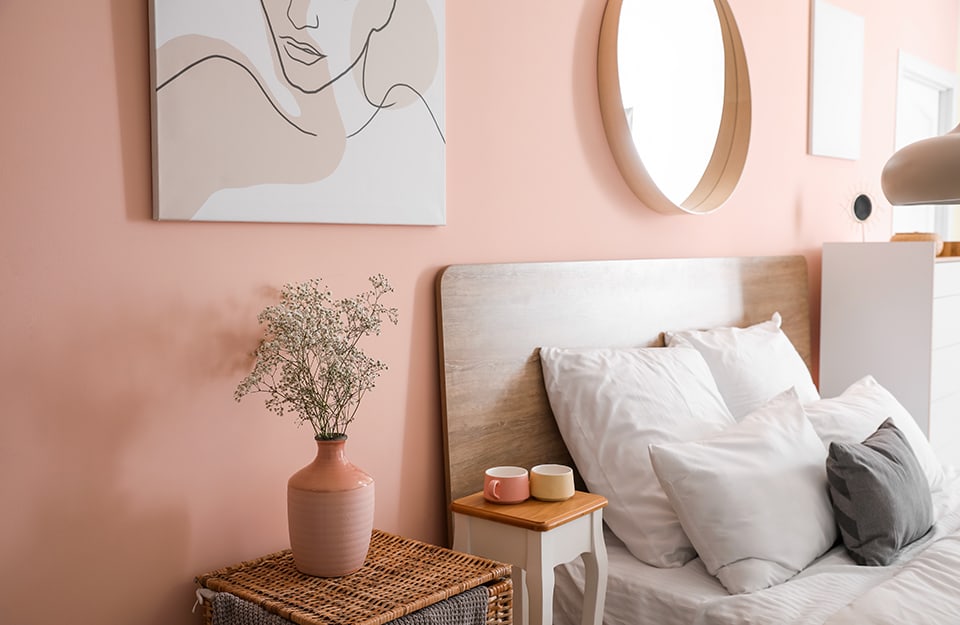 Detail of a bedroom with a pink wall with hanging pictures and a circular mirror above the bed. The headboard is thin, made of wood and with minimal lines. Next to the bed a small wooden table with cups on it, and another wicker table with a pink vase with flowers on it. On the opposite side of the bed is a tall, white chest of drawers;