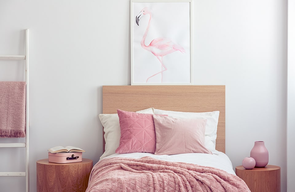 A bedroom in shades of white and pink, with a wooden bed with pink blankets and pillows, two cylindrical wooden bedside tables with pink jars on top and a small metal chest, also pink, as well as an open book. Above the bed is a print of a red flamingo, framed in white, and to the left a white ladder used as a coat rack;