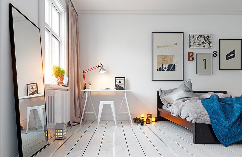 An industrial-style bedroom in shades of white, with parquet flooring and white walls. A large mirror leaning against the wall reflects part of the room. The bed has a basic black wooden frame and next to it is a white desk on trestles, with a stool that is also white. On the desk are a study lamp, a photo and office objects. On the floor are several lit candles. Hanging on the wall, above the bed, are several prints with typefaces