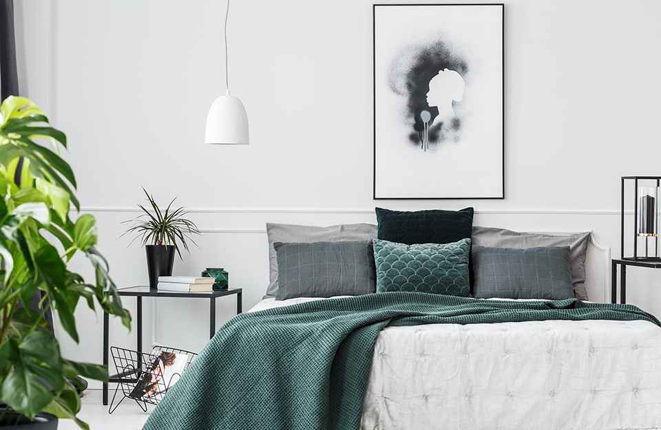 Modern bedroom in shades of white, grey and green. The wall behind the bed is white, with a painting of a woman's profile hanging on it. The bed has pillows and blankets in green and grey. On one side is a black metal bedside table with books, a seedling and a jar on top and a magazine rack also in black metal underneath. On the other side is a twin table with a square black lamp on top. A large monstera plant can be glimpsed on the left. A small white dome-shaped chandelier descends from the ceiling;