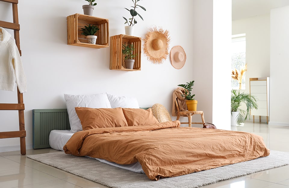 Boho-chic style bedroom with white walls, a bed resting directly on the floor, on top of a rug, and behind an old shutter painted green to act as a headboard. To one side of the bed is a wooden ladder that serves as a clothes rack. On the other side is an old stuffed chair on which there are clothes and a plant in a yellow pot. Above the bed, fruit boxes serve as shelves and contain plants. Hanging on the wall are also two straw hats. Another room can be glimpsed beyond a column;