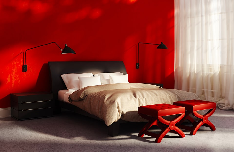 Bedroom with deep red walls and grey floor. A semi-transparent white curtain covers the window. Two black directional lamps hang on the wall and at the foot of the bed are two red quilted and lacquered stools;