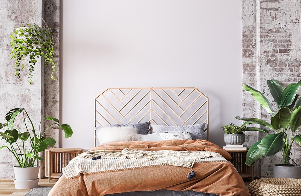 Rustic-modern style bedroom with a white wall between columns and walls with exposed bricks. The floor is parquet and the bed has a wooden frame with an open headboard with a herringbone pattern. On the bed are orange blankets and white and grey pillows. On either side are circular wooden bedside tables. Above one of them are books and a plant in an antique-looking pot. In the rest of the room there are a couple of plants in pots on the floor and a wall cabinet;