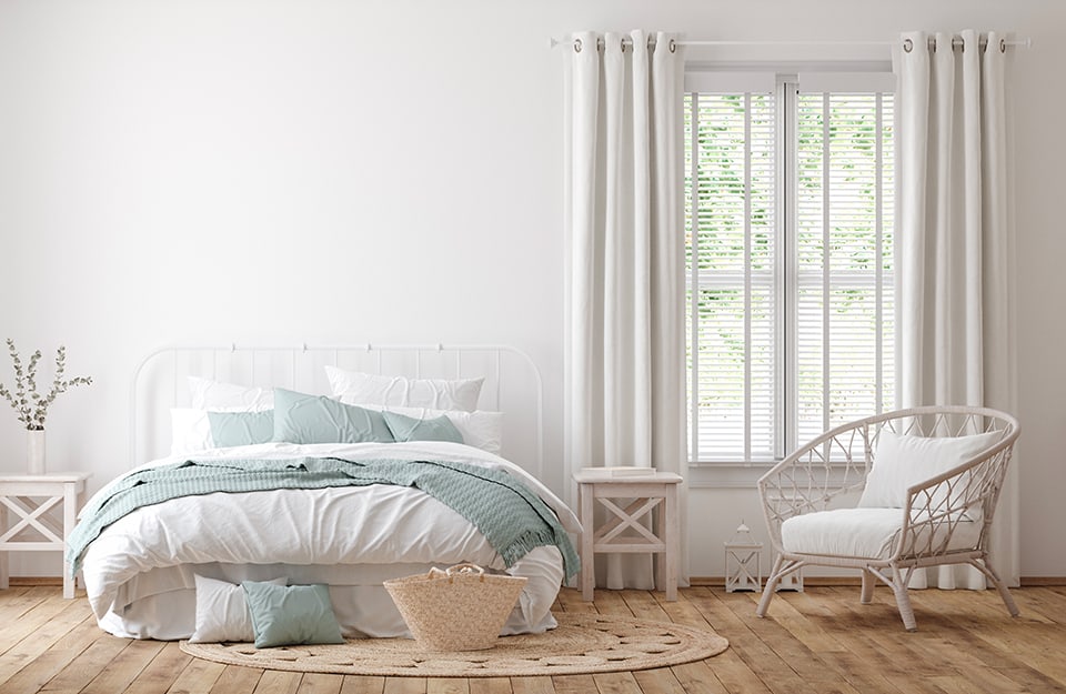 Rustic chic bedroom. The room has all-white walls and a parquet floor. A window with white curtains overlooks what looks like a garden. The bed has a minimal metal frame and on either side are rustic white bedside tables. Above one of them is a tall, thin, white vase with twigs inside. On the floor is a circular rug with cushions and a wicker basket on top. In front of the window is a rustic white armchair;