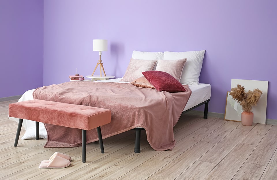 A very bare bedroom with a purple wall. The bed is on a simple black metal frame. Blankets and pillow are on white and pink. On one side of the bed a small white circular table with a lamp on it and another smaller table with books and candles. On the other side, frames resting on the wall and a pink vase with dried flowers inside. The floor is light parquet and pink slippers can be seen;