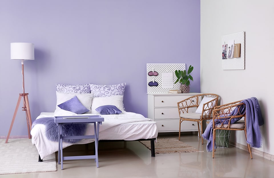 Bedroom in a modern, minimalist style with a light purple wall. The bed is on a simple black metal frame. The blankets and pillows are on white and purple. At one side of the bed, above a white carpet, a lamp with a wooden tripod. At the end of the bed a small purple table. On the other side of the bed a smaller rug, a white chest of drawers with books and a plant on it, and two bamboo chairs. On the walls hang two white storage panels: on one there are books and prints, on the other there are night masks and a calendar;