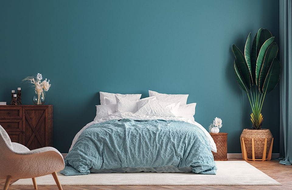 Dormitorio en tonos azul claro, con una cama de matrimonio con ropa de cama azul claro y blanca. Varias cestas de mimbre esparcidas por la habitación, una de ellas es un macetero con una planta grande dentro. El suelo es de parqué y se ve, a la izquierda, parte de un sillón, y un mueble de madera con jarrones y chucherías encima. La pared de detrás de la cama es de color azul claro;