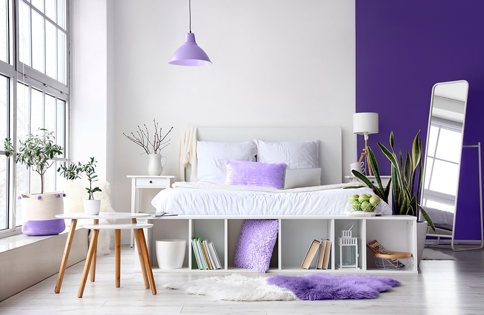 Bright and spacious room with a large window and a windowsill with a purple and white vase holding a plant. The parquet floor is white. The wall behind the bed is white and then turns purple. The bed is white painted wood and at the bottom there is a low horizontal shelf containing books and a purple cushion. There are more purple cushions in the room, and on the floor is a long two-coloured carpet in white and purple. On either side of the bed are white bedside tables with a drawer, and above them a lamp and a vase. A purple, industrial-style chandelier descends from the ceiling. To the right of the bed is a plant in a white pot. Just beyond it is a large metal-framed table;