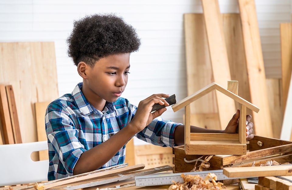 Bambino in un laboratorio di falegnameria sta carteggiando una casetta di legno in miniatura