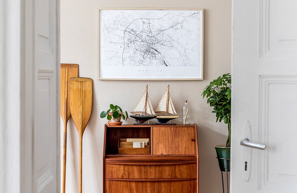 Entrada con una consola-escritorio de madera natural, con maquetas de veleros sobre ella, un cuadro con un mapa y remos de canoa al lado. Dos puertas blancas abiertas enmarcan la escena;