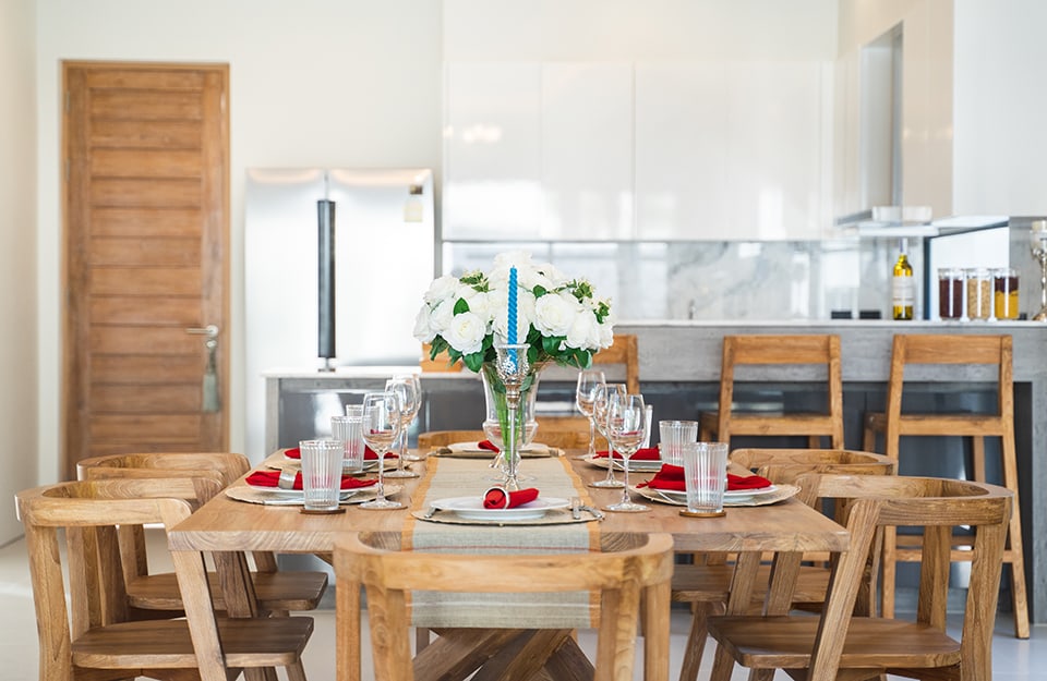 Mesa de comedor decorada festivamente en madera natural. Detrás se ve una puerta de madera natural y una cocina en tonos blancos y grises;