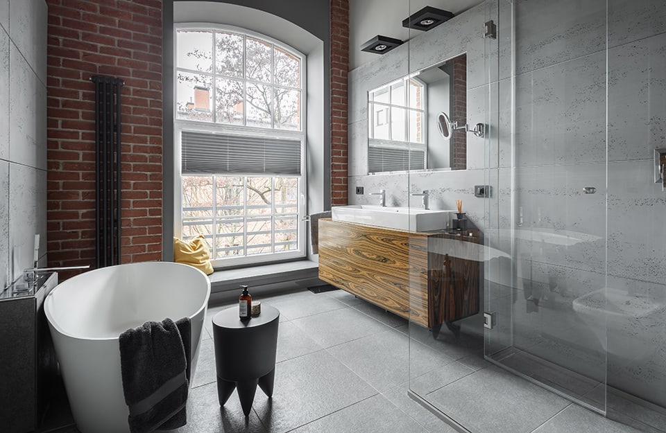 Industrial-style bathroom in a room with rough concrete walls and another wall with exposed bricks. The shower is entirely transparent and there is a white bathtub in minimal style. The washbasin-cabinet is made of natural wood with very obvious grain