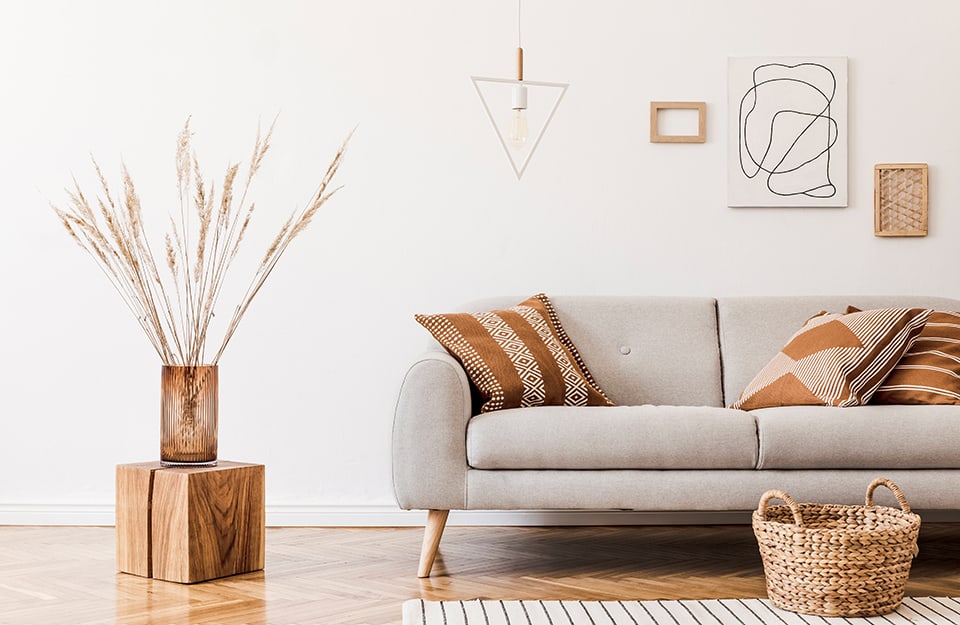 A Scandinavian-style living room with a grey sofa, brown cushions with ethnic geometric patterns, a wicker basket on the floor, squares on the walls and a natural wood cube that serves as a coffee table or stool;