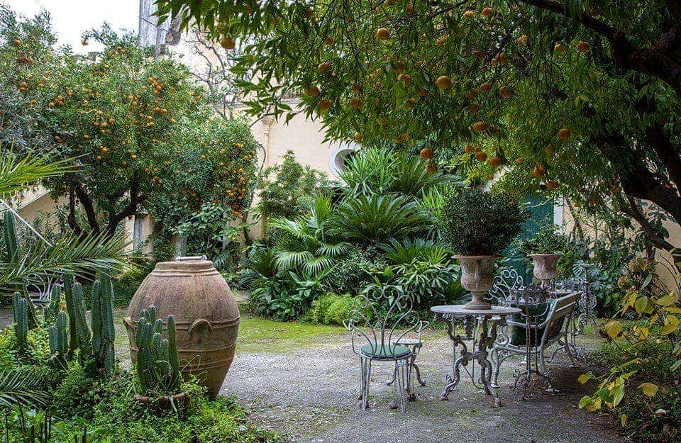 Un cortile in stile mediterraneo con molte piante, anche di agrumi, grandi orci e sedie e tavolo in metallo