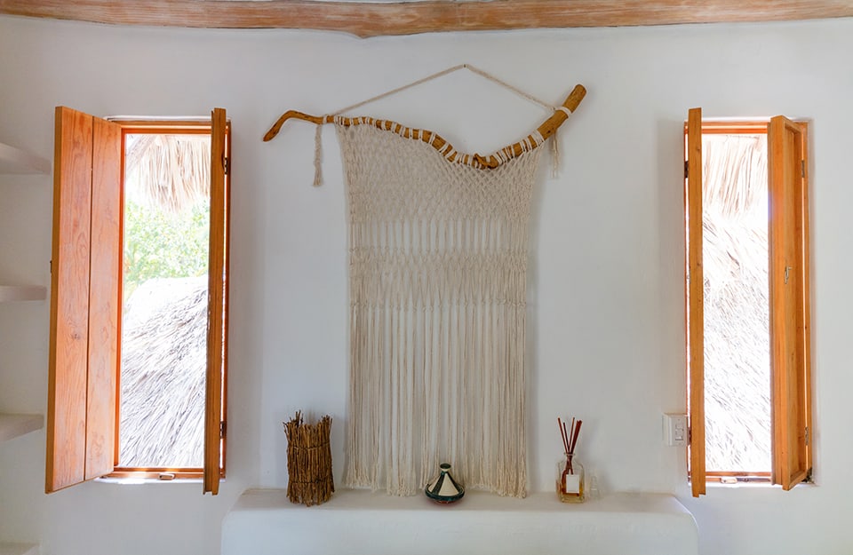 Una decorazione realizzato con un ramo di legno ritorno e tessuto è la protagonista di una parete in una stanza bianca con due finestre e una mensola con soprammobili in stile etnico