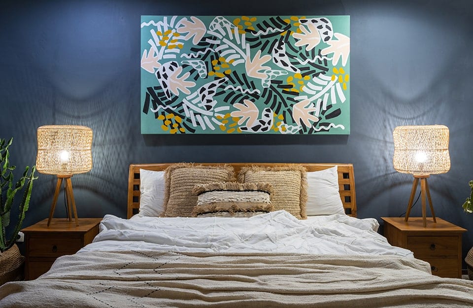 Camera da letto con parete blu pastello su cui è appeso, sopra la testiera, un quadro con motivi botanici colorati. Due abat-jour in legno intrecciato sono ai due lati del letto, sopra dei comodini