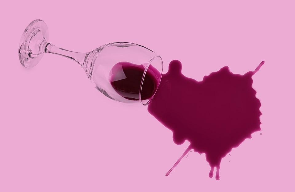 Un bicchiere di vino bordeaux rovesciato con vino che fuoriesce e crea una macchia, su fondo rosa
