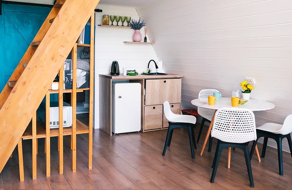 Una piccola cucina in una mansarda, con tavolo da pranzo, lavello, frigorifero e piccola scala in legno che funge anche da libreria