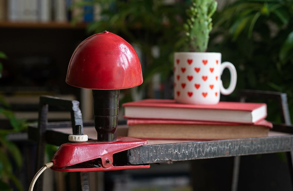 Dettaglio di uno studio con una lampada vintage a fungo con clip da agganciare al tavolo, sul quale ci sono dei libri e una tazza bianca con dei cuori usata come vaso per una piantina