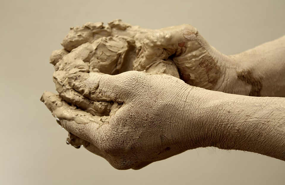 Delle mani infangate stanno manipolando una palla di fango, su sfondo monocromatico color fango