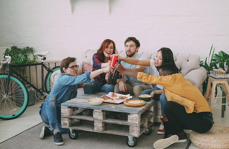 Un gruppo di amici o di coinquilini cena insieme con pizza e pop-corn e brinda sopra un tavolino costruito con un pallet riciclato, in un salotto moderno open space