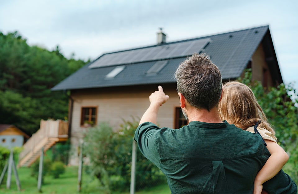 Un padre tiene in braccio una figlia nel giardino di casa. Sono visti da dietro e il padre indica con il dito i pannelli solari sul tetto della casa