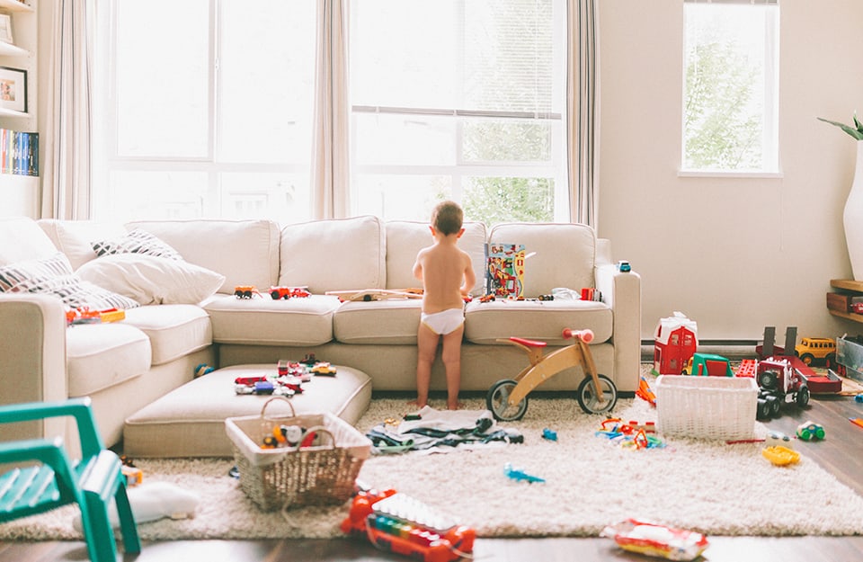 Un bambino piccolo col pannolino visto di spalle in piedi davanti al sofà, in un salotto molto disordinato pieno di giocattoli sparsi ovunque