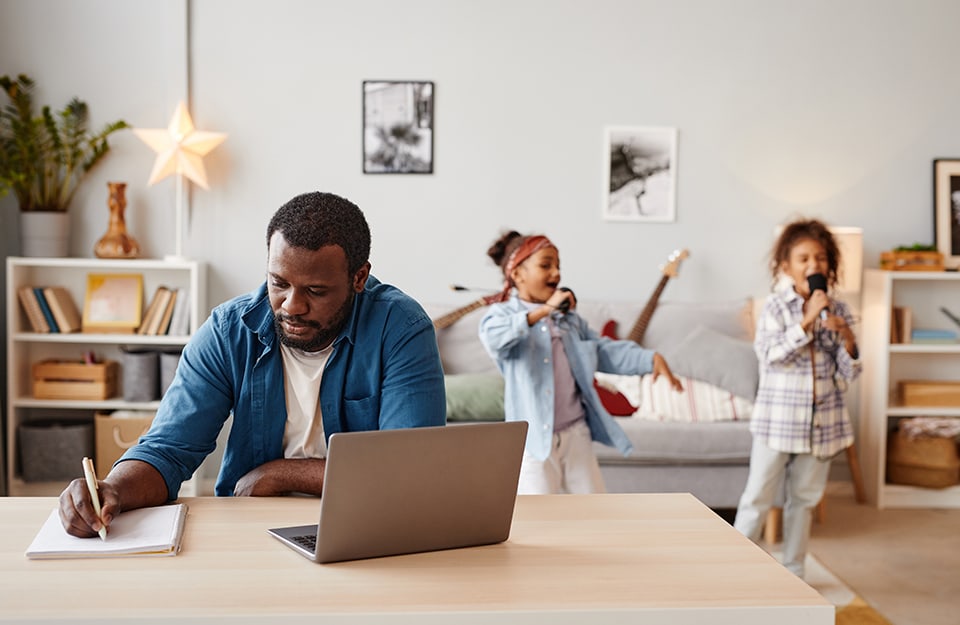 Un uomo sta cercando di lavorare con laptop e blocco note nel salotto di casa mentre sullo sfondo le due figlie giocano a cantare al karaoke