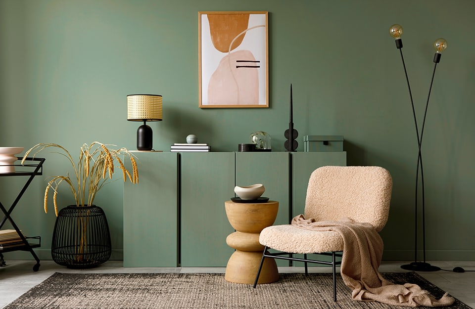 Una salotto tutto sui toni del verde salvi, pareti comprese, con elementi verticali sui quali sono in mostra soprammobili e una lampada. Ci sono inoltre una poltrona pelosa rosa, un tavolino in legno a forma di clessidra, un tappeto e una pianta