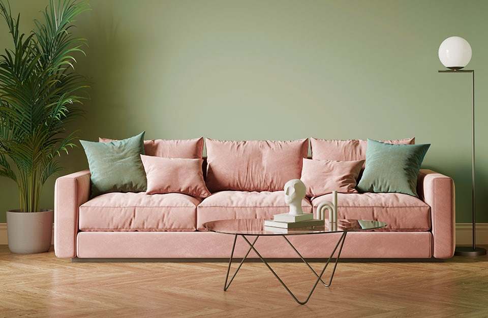 Un salotto con pareti verdi salvia, un sofà a tre posti rosa pastello con cuscini rosa e verdi, una lampada da terra, un tavolino minimalista in metallo e una pianta in vaso, su un parquet