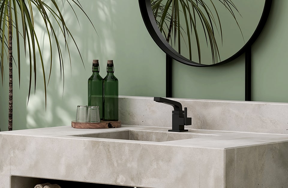 Primo piano di tre quarti a un lavandino squadrato color cemento in un bagno con pareti verde salvia e uno specchio che riflette parzialmente una pianta