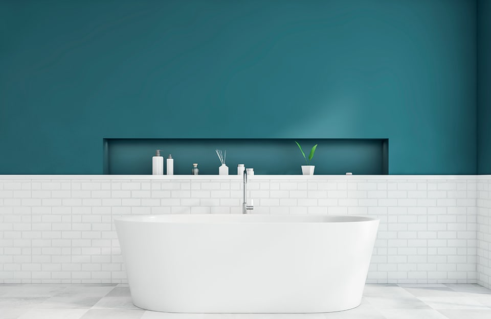 Un bagno con parete color ottanio con nicchia in cui sono disposti saponi ed essenze, muretto in piastrelle bianche e grande vasca da bagno bianca
