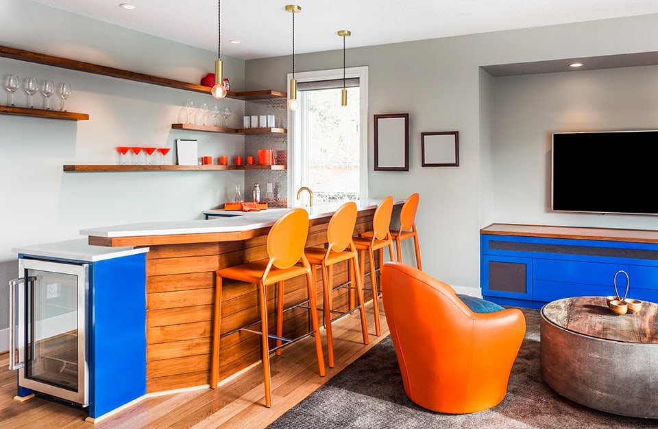 L'angolo bar con bancone in legno, sgabelli arancioni, mini-frigo blu, scaffali, lampadari a sospensione sopra il bancone e un salottino nei pressi del bar
