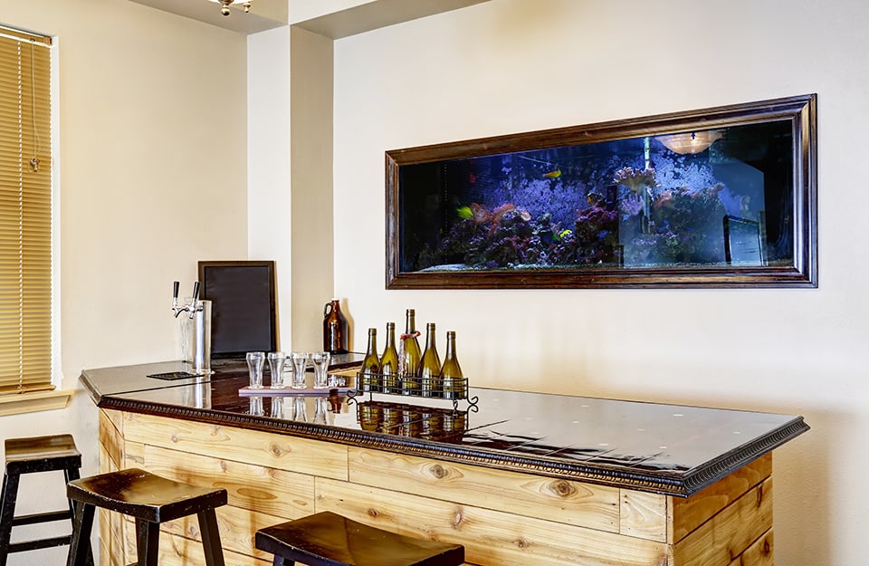Un angolo bar domestico con bancone in legno rustico e acquario orizzontale a muro dietro al bancone