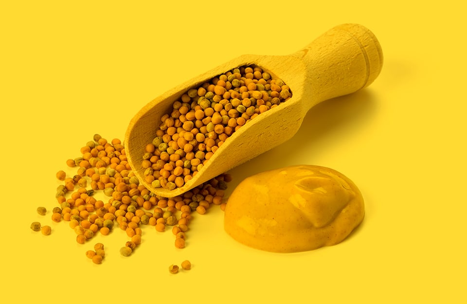 Un cucchiaio di erboristeria pieno di semi di senape, che fuoriescono anche sulla superficie, accanto a della salsa di senape, su sfondo giallo senape