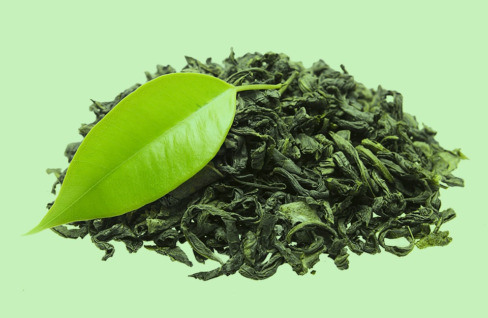 Delle foglie essiccate e sminuzzate di tè e una foglia integra, su sfondo color tè verde