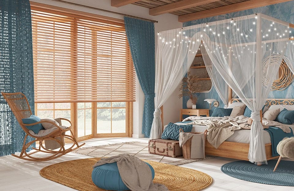 Grande camera da letto affacciata sul mare, in stile coastal, con pareti azzurre, letto a baldacchino con tende bianche semitrasparenti, tappeti in tessuto grezzo, poltrona in vimini e grande uso del legno naturale