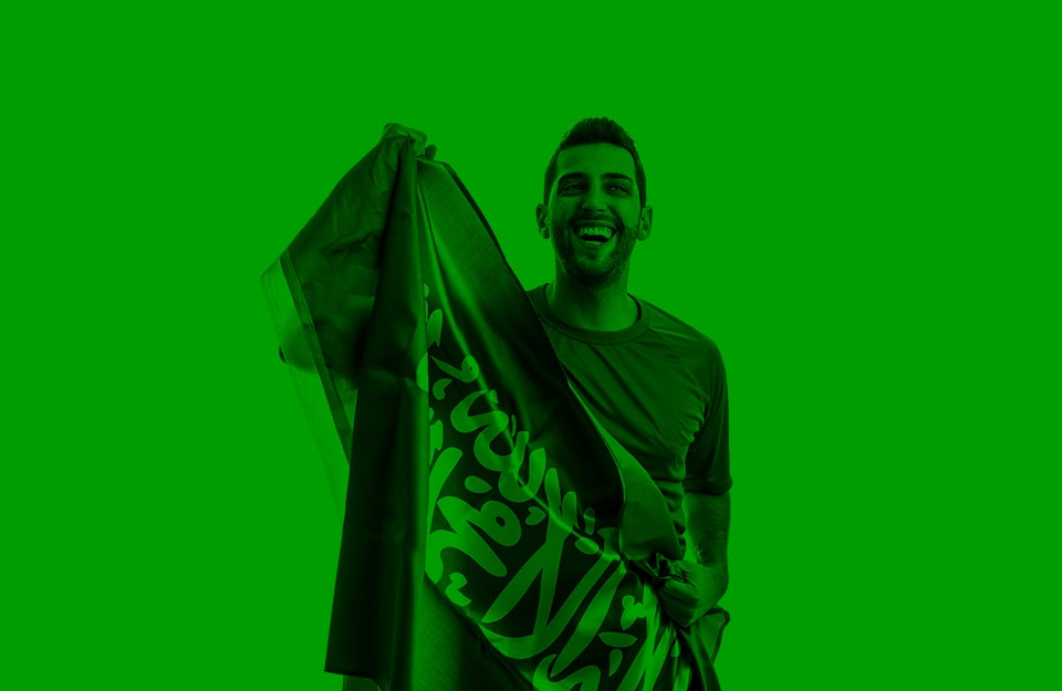 Un ragazzo sorridente tiene in mano la bandiera dell'Arabia Saudita e l'immagine è tutta virata sui toni del verde