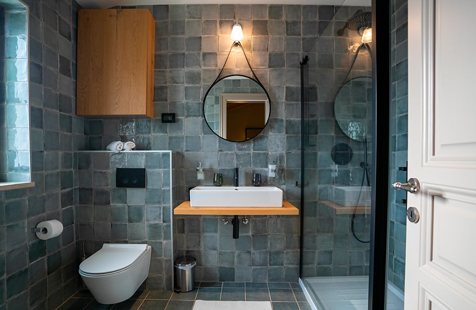 Bagno completamente piastrellato con piastrelle sui toni dell'azzurro-grigio, specchio circolare, lavandino su mensola, box doccia e sanitari bianchi
