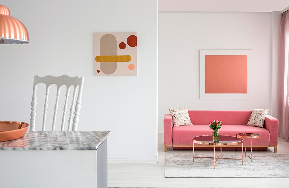 Dettaglio di una cucina affacciata su un salotto, tutto sui toni del bianco e del rosa, con dettagli rosa metallizzato come i tavolini da caffè e il lampadario della cucina