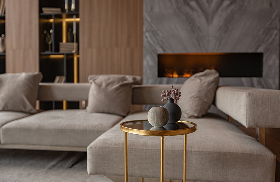 Salotto moderni con grandi sofà squadrati color tortora, caminetto geometrico orizzontale e tavolino da caffè circolare metallizzato color oro bruno