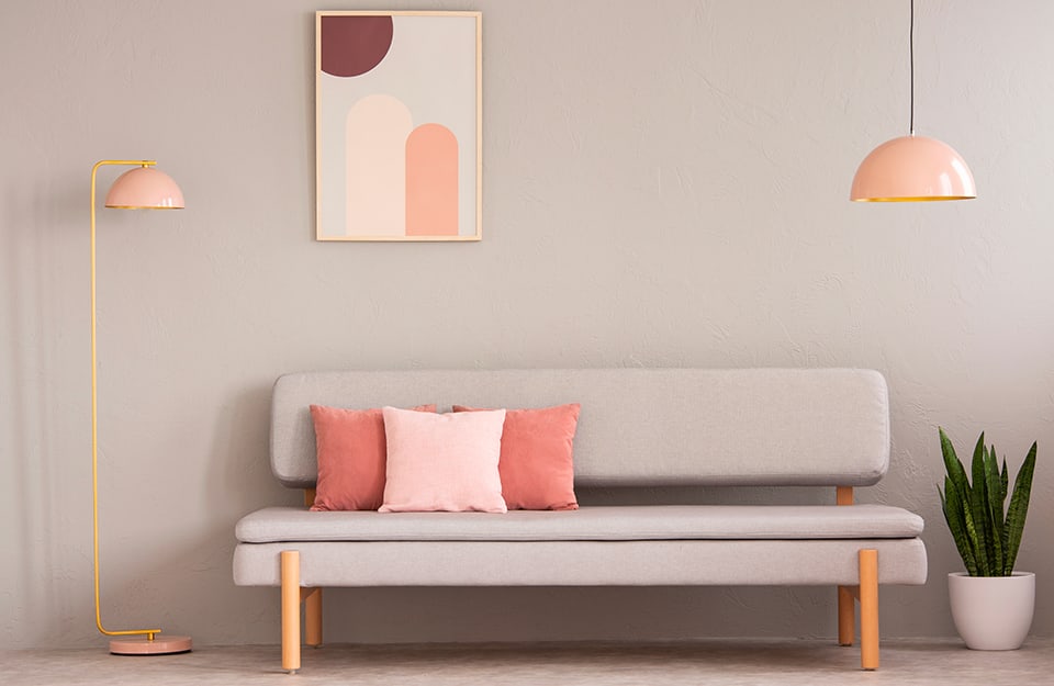 Angolo di un salotto o di un ingresso, con panca di colore chiaro, della stessa tinta della parete, e cuscini di varie tinte di rosa. Una lampada a soffitto e una da terra, entrambe tondeggianti, sono rosa, così come alcuni dettagli di un quadro geometrico astratto