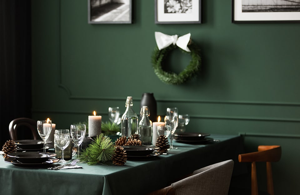 Vista ravvicinata e angolata su un tavolo da pranzo addobbato per le feste natalizia, con tovaglia verde smeraldo scuro che richiama il colore delle pareti, dove si intravedono quadri e una corona di foglie decorativa