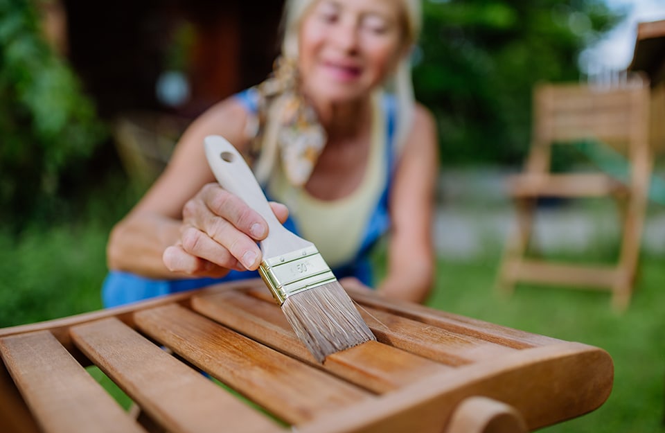 Una donna, vista fuori fuoco, sta dipingendo una sedia da giardino in legno con una finitura all'acqua trasparente